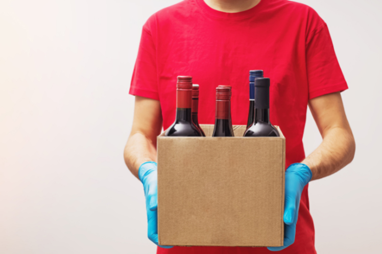 A delivery personnel wearing gloves and holding a box filled with wine bottles