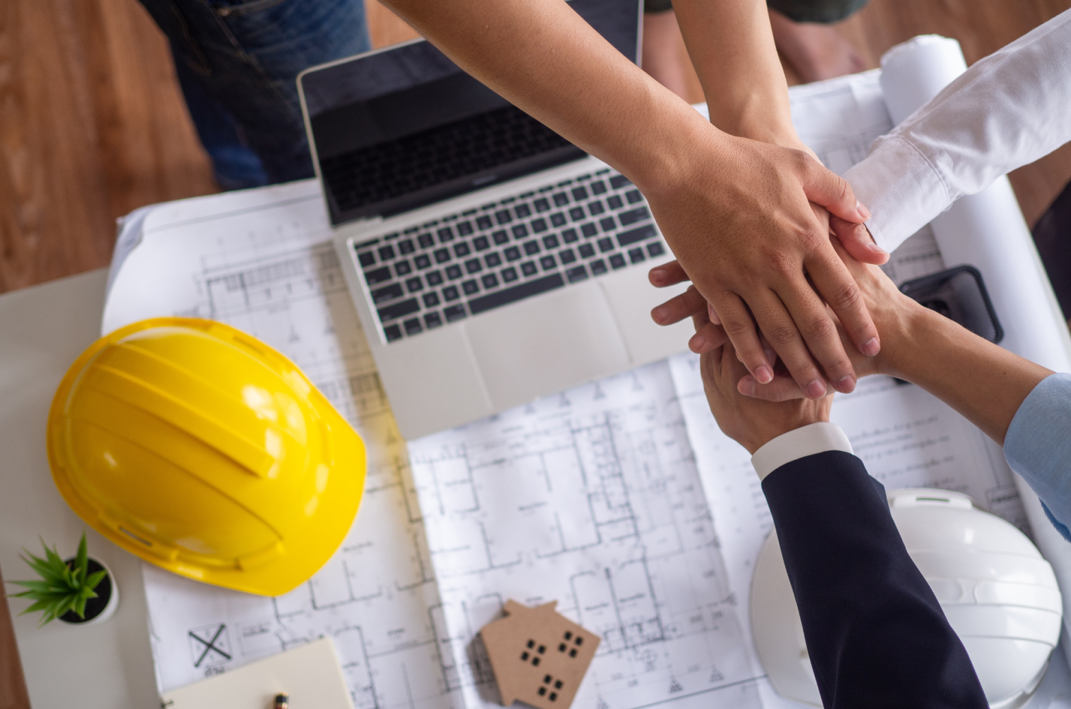Three people doing a team hand gesture over a blue print, laptop, and construction hats