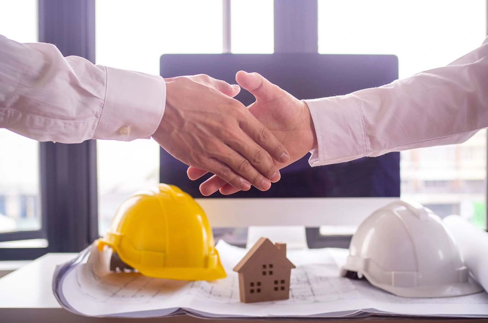 Two people shaking hands with construction blueprints, a notebook, a hard hat, and a small wooden house behind them