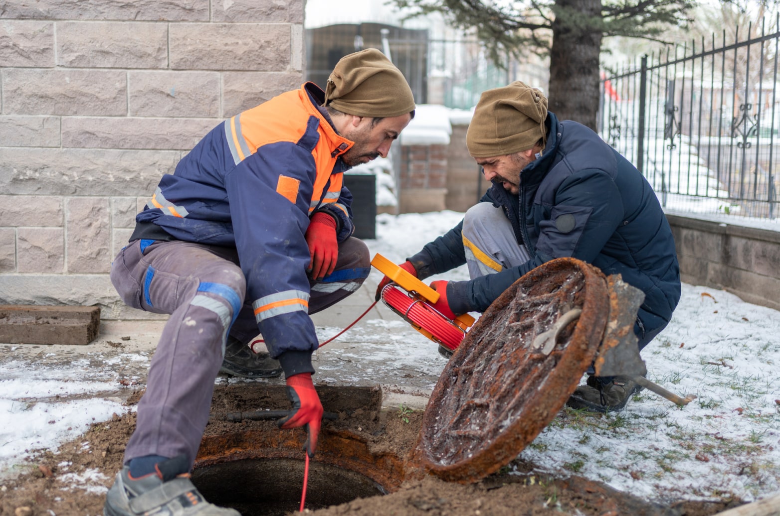 Two contractors in cold-weather gear fishing a line into a sewer