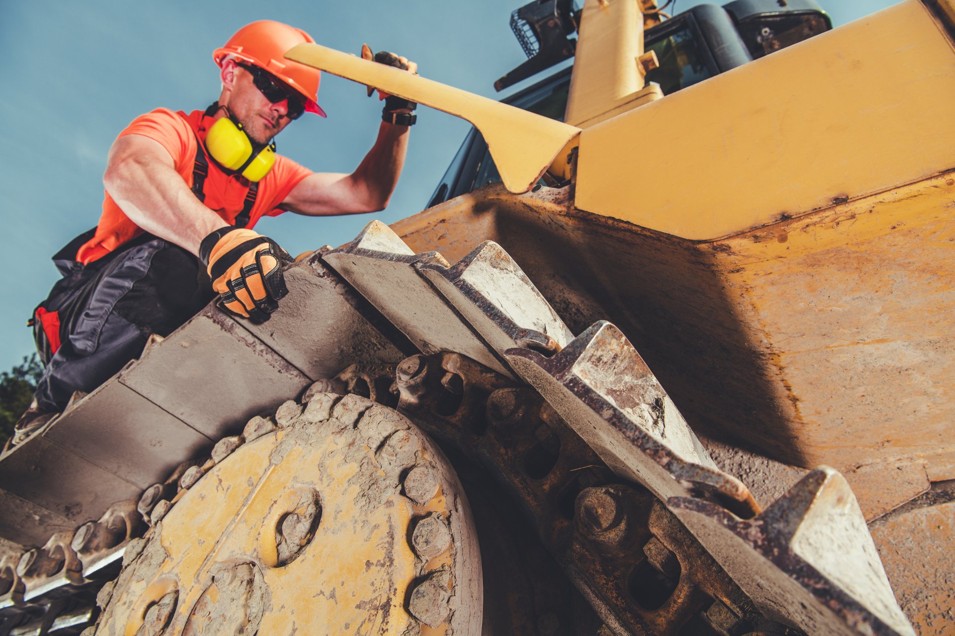 Heavy machinery operator wearing an orange shirt, hard hat, and sunglasses checking machinery tracks