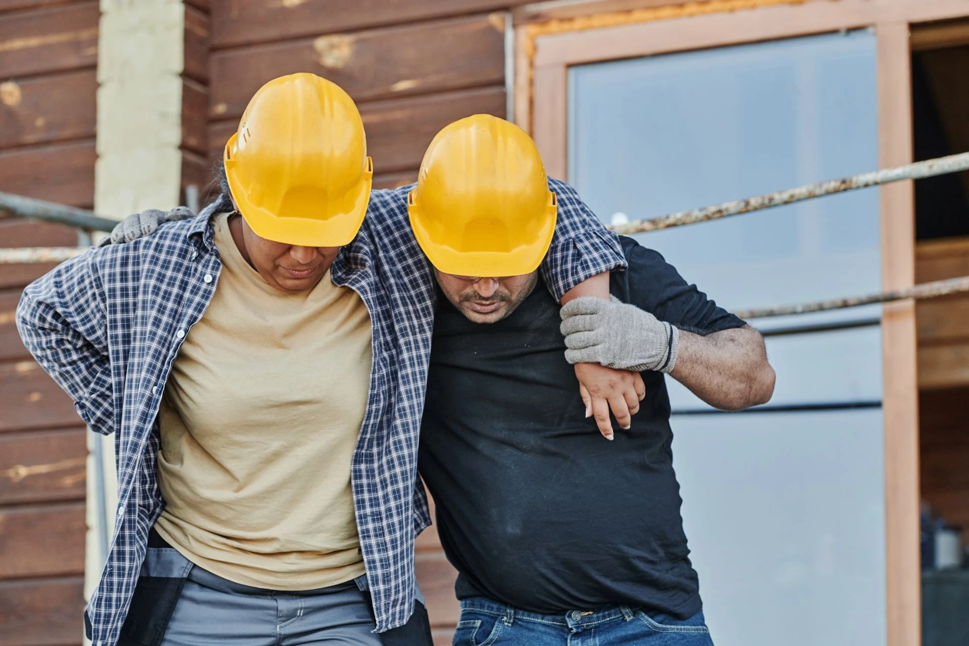Two construction workers with yellow hard hats walk together, one supporting the other who appears to have a back injury