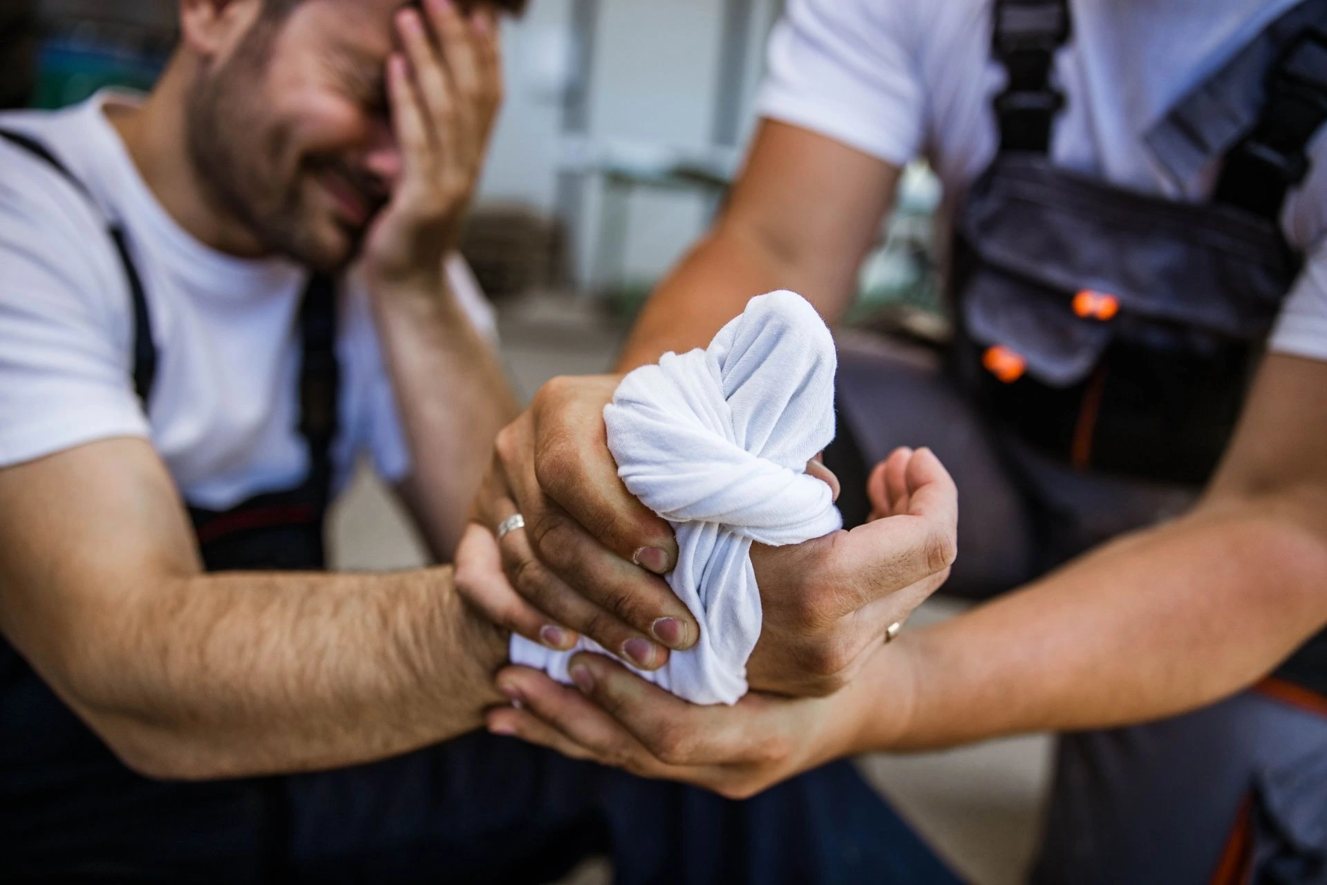An injured worker with his thumb wrapped in a makeshift bandage is supported by a coworker.