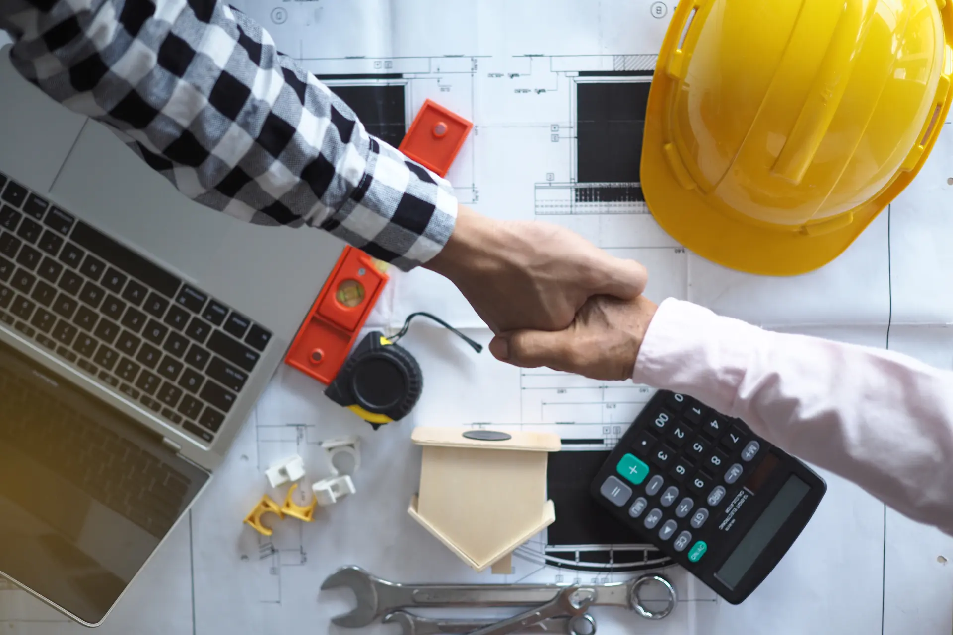 A contractor and a client shaking hands over blueprints and construction tools
