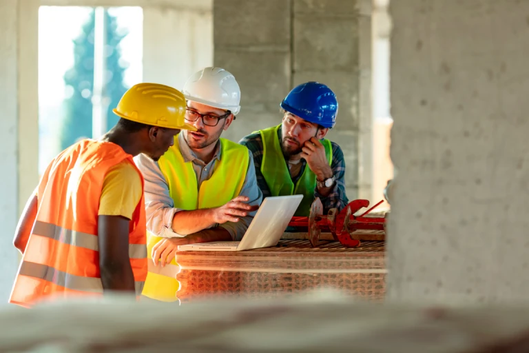 Three construction workers wearing safety vests and hard hats review project plans on a laptop at an active building site
