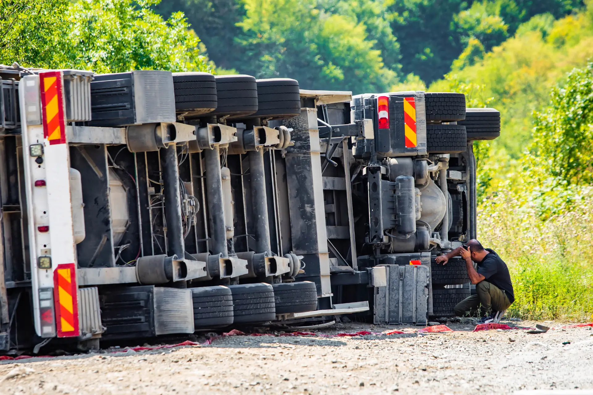 A cargo truck overturned on the road due to an accident, a driver leaning down beside it
