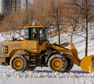 Yellow front-end loader clearing snow in a city park during winter.